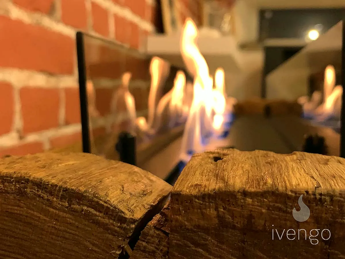Пламя биокамина iVENGO за стеклом у кирпичной стены, впереди срезы брёвен